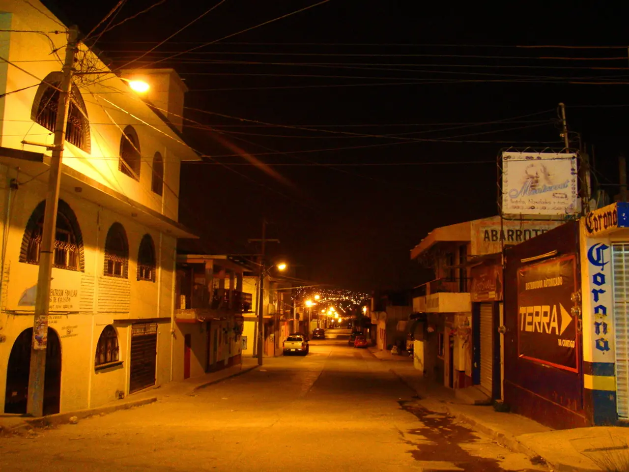 In this image on both sides there are buildings, electric poles. There are few vehicles parked in...