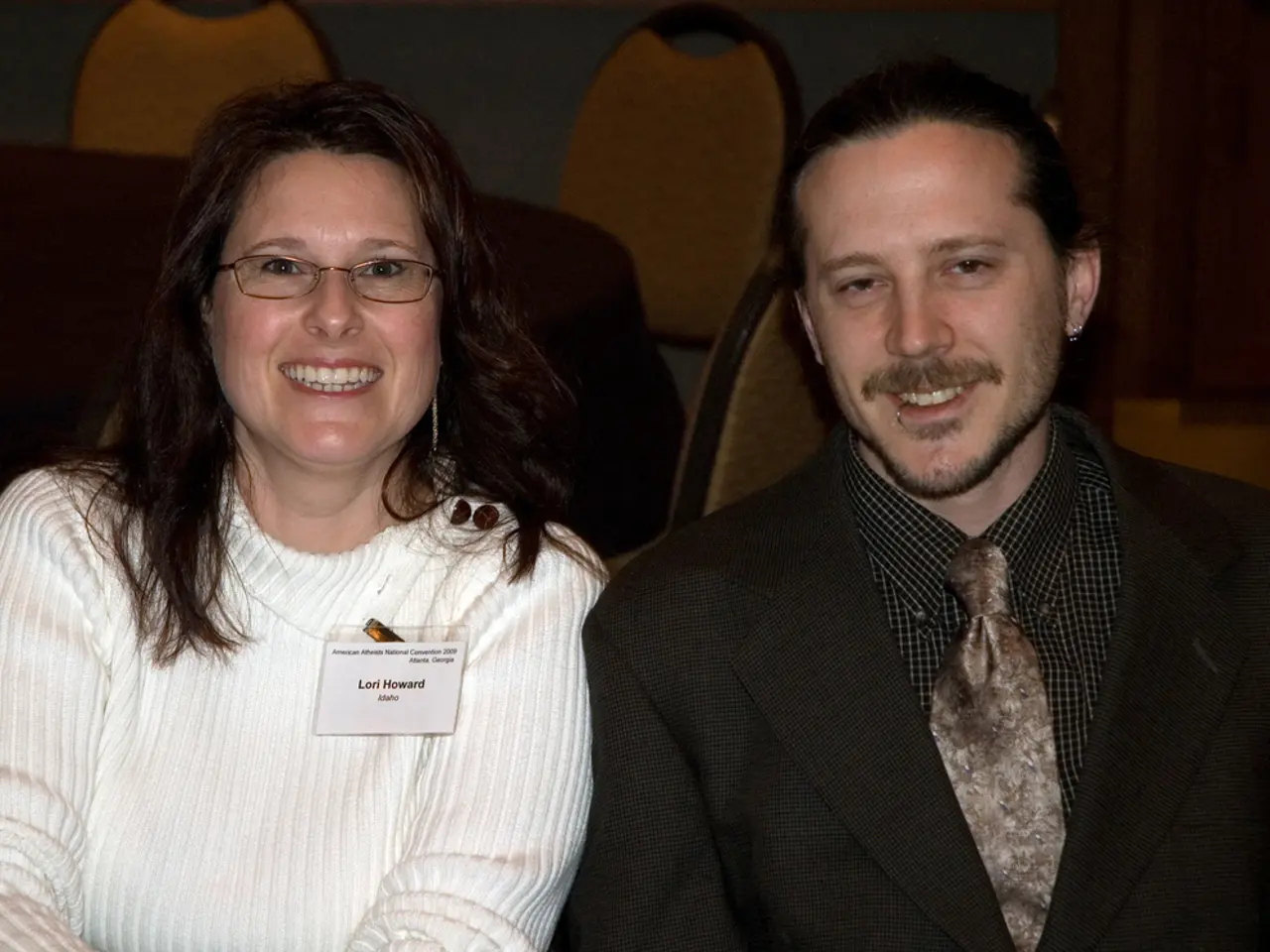 There is a man and a woman. Woman is wearing a specs and a badge. In the back there are chairs.