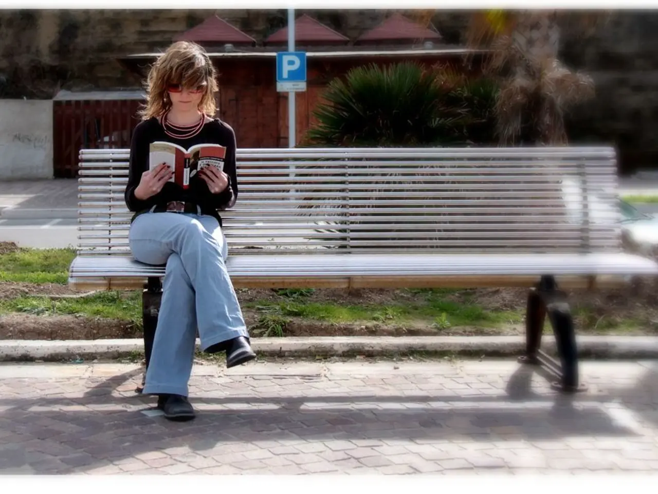 A woman is sitting on a bench. She wears a T shirt and jeans pant. She is reading a book. She wears...