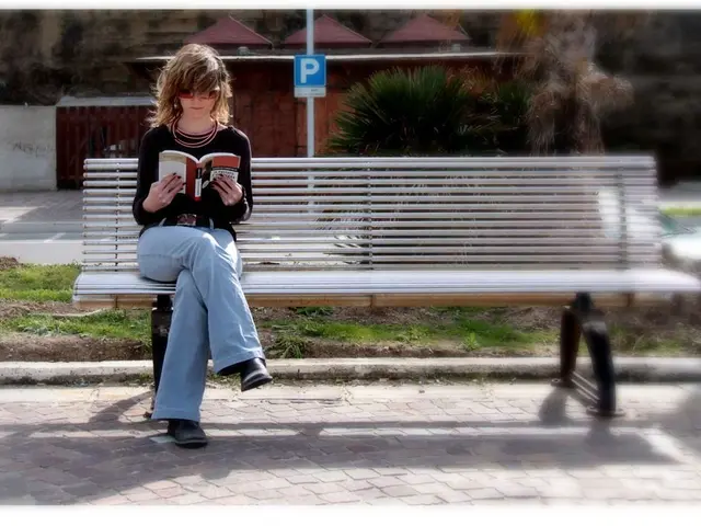 A woman is sitting on a bench. She wears a T shirt and jeans pant. She is reading a book. She wears...