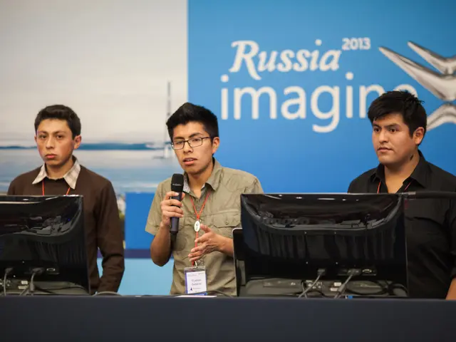 In the picture we can see three boys standing near the desk on it, we can see two computer systems...