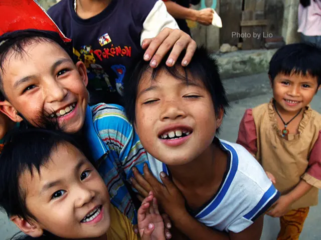 In this image we can see kids standing and smiling. In the background there is a road and a wall.