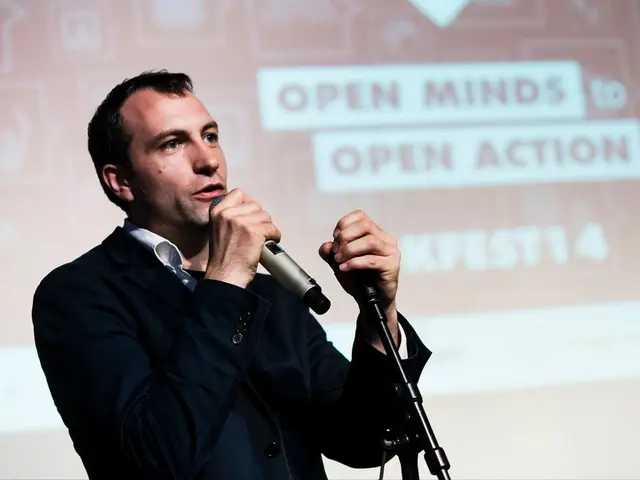 In this picture a man is speaking with the help of microphone, in the background we can see a...